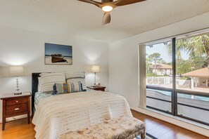 Un dormitorio con una cama, dos mesitas de noche, un ventilador de techo, un ventanal con vistas a una piscina y palmeras, y un cuadro de un barco en la pared.