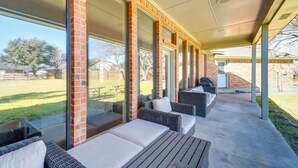 A covered outdoor seating area with wicker furniture and cushions, large windows, and a view of a grassy yard. - ~ 3 Mi to Dtwn: Aubrey Vacation Rental w/ Patio! (Aubrey)