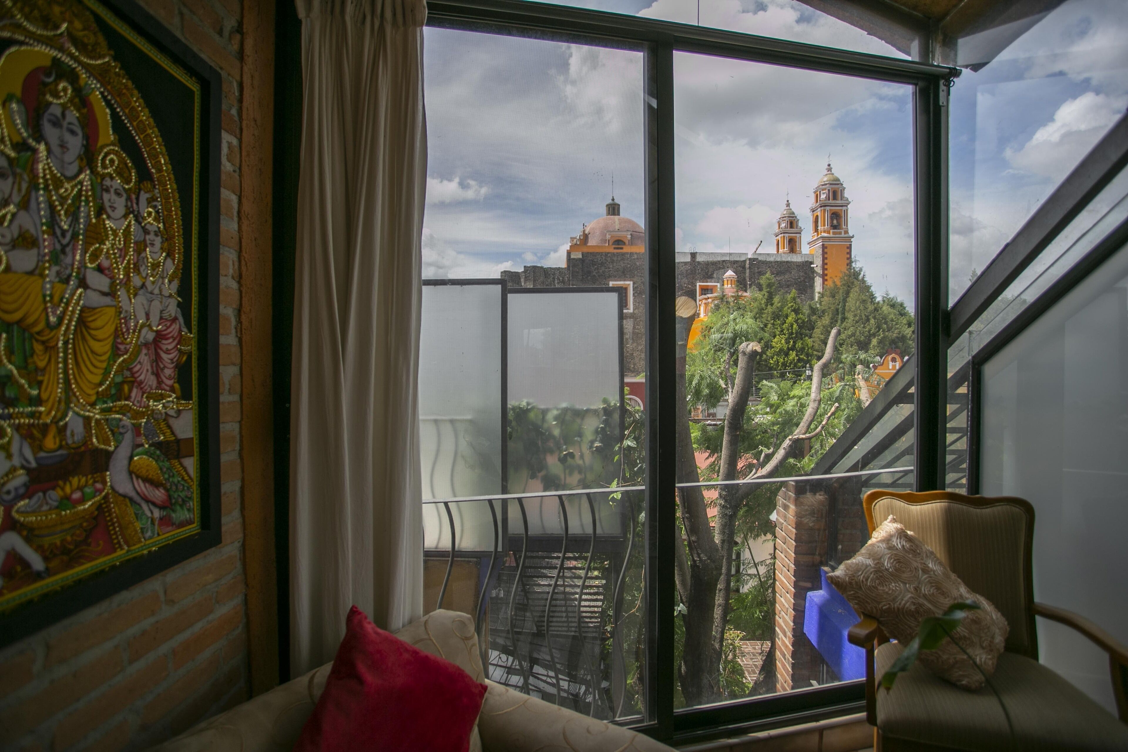 Una habitación con un amplio ventanal que da a un paisaje urbano, una obra de arte enmarcada en la pared y una silla con un peluche.