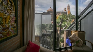 A room with a large window overlooking a cityscape, a framed artwork on the wall, and a chair with a stuffed toy. - Martinlandia  (Puebla)