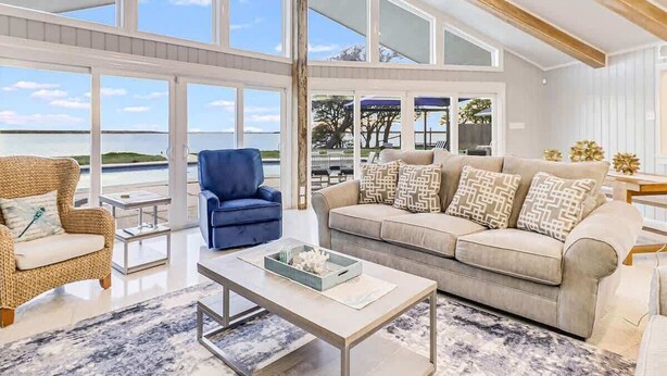 A spacious living area with large windows, a blue armchair, a beige sofa, and a coffee table. - Whispering Breezes at Windy Hill (Ingleside)
