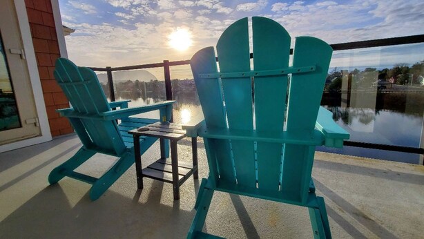 A balcony with two turquoise Adirondack chairs and a small table, overlooking a body of water and a cityscape. - BP 410 River Song Condo.  3 blocks from the beach (Seaside)