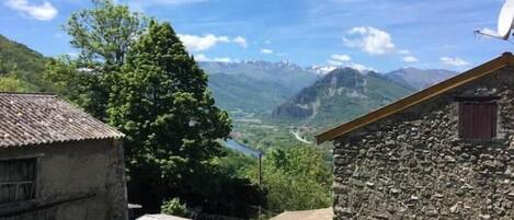 A rustic stone building with a corrugated metal roof, a small adjacent shed, and a large tree in a mountainous landscape.
