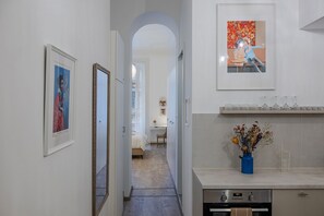 Un pasillo con obras de arte enmarcadas, una cama, un escritorio y una zona de cocina con un jarrón de flores.