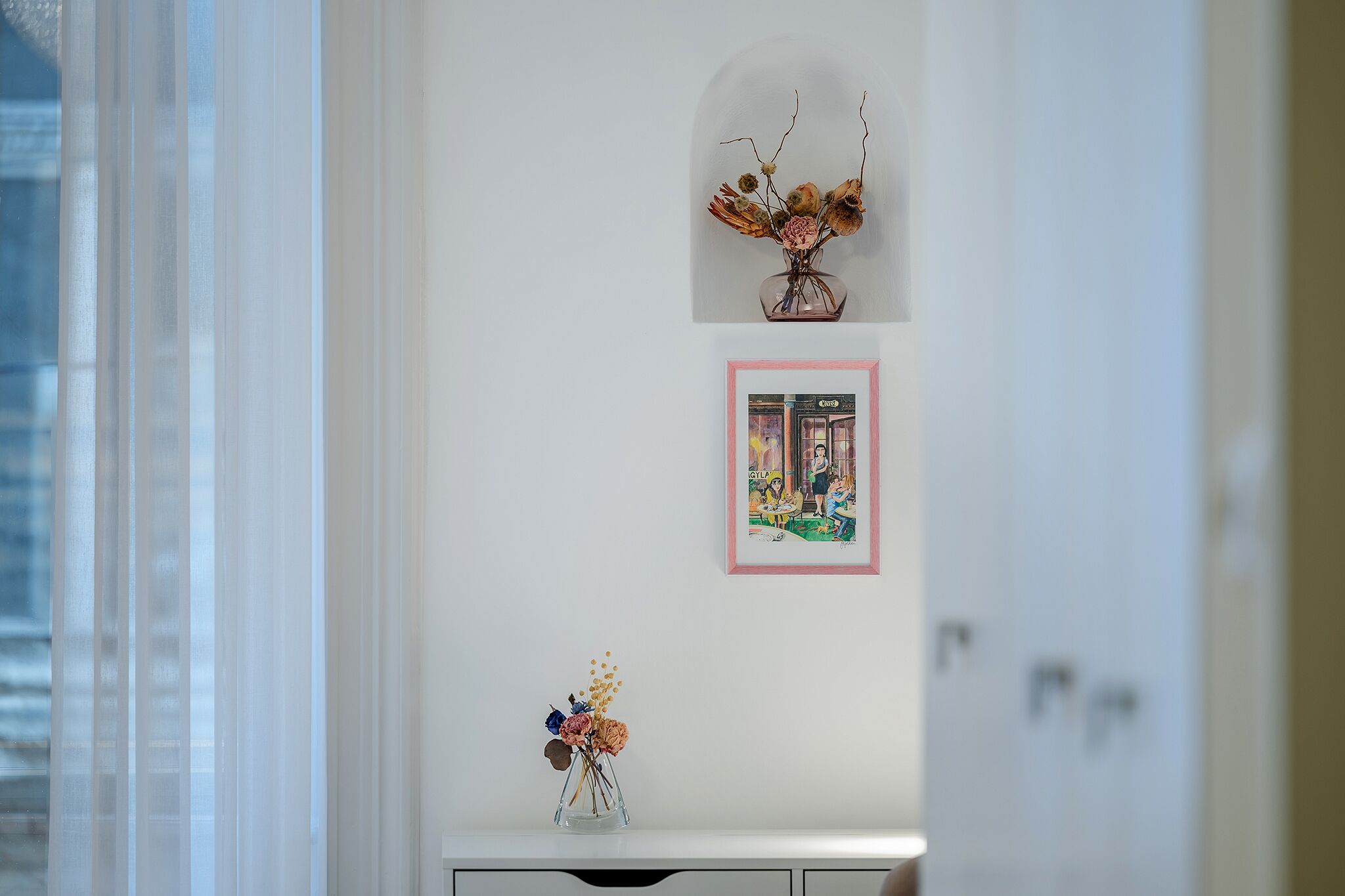 A white wall with three framed items: a vase with dried flowers, a framed picture of a street scene, and a smaller framed picture with a pink border.