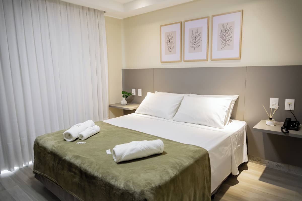 Une chambre d’hôtel avec un lit soigneusement fait, une couverture verte, des oreillers blancs, une table de chevet avec une plante et, au mur, des œuvres d’art représentant des feuilles, encadrées.