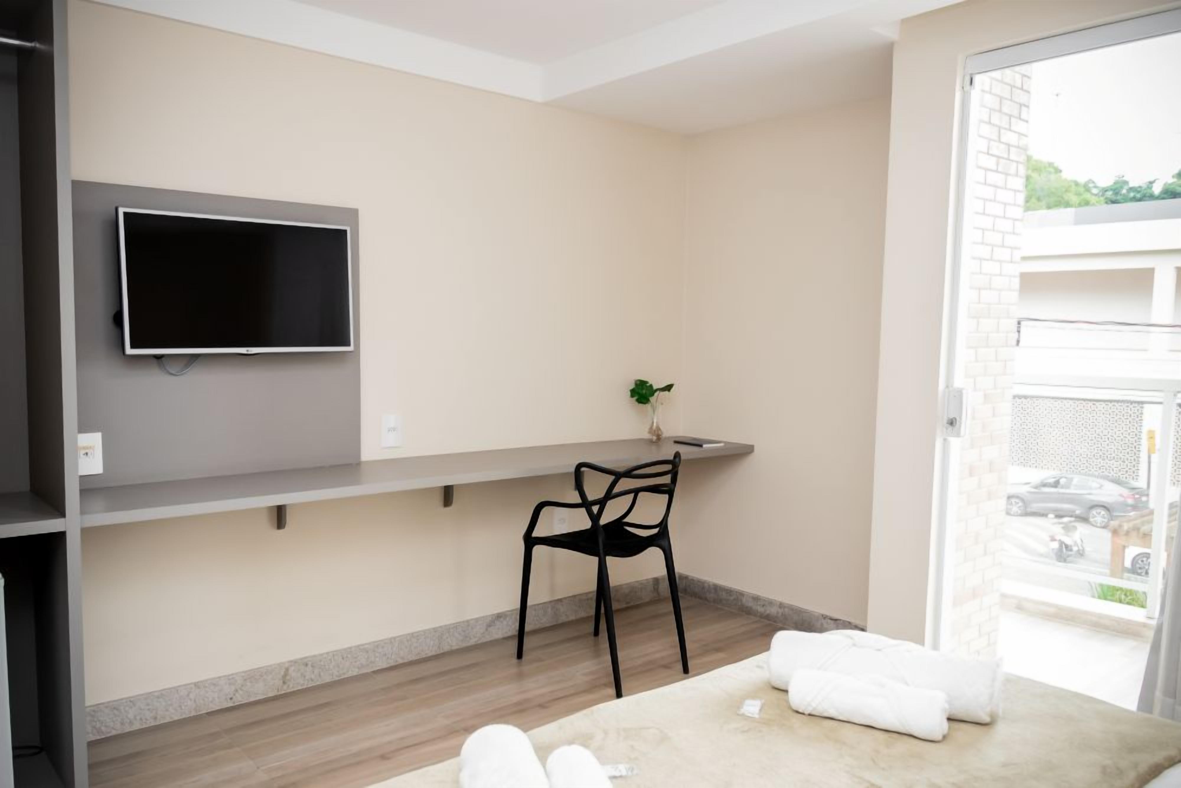 A hotel room with a flat-screen TV mounted on the wall, a small table with a chair, and a window with a view of the outside.