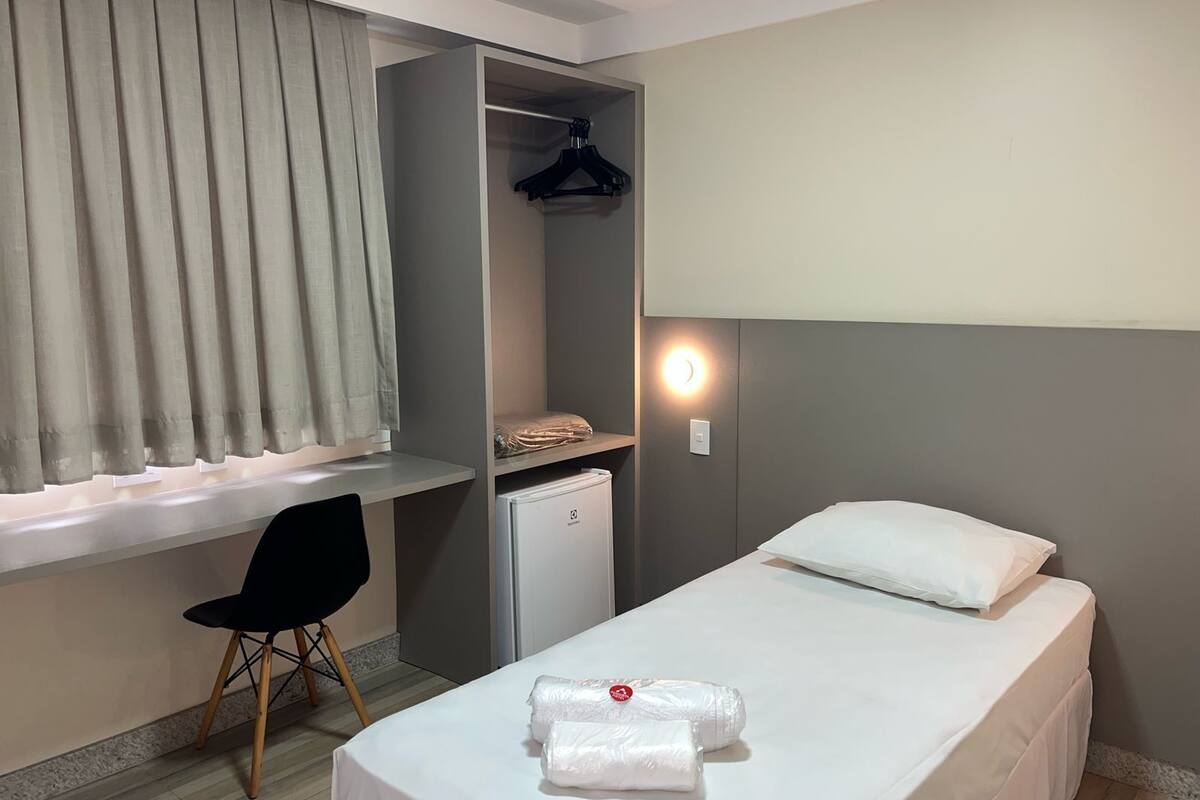 Une chambre d’hôtel de taille réduite, comprenant un lit simple, un petit bureau, une chaise, une armoire et un mini-réfrigérateur.
