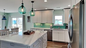 A modern kitchen with white cabinets, a large island, and a stainless steel refrigerator. - The Salty Seahorse | Across from Beach + Views! (North Topsail Beach)