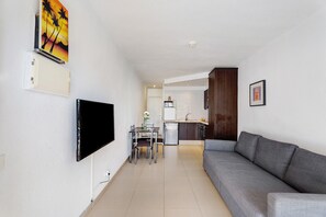 A modern living room with a flat-screen TV, a grey sofa, a dining table with chairs, and a kitchen in the background.