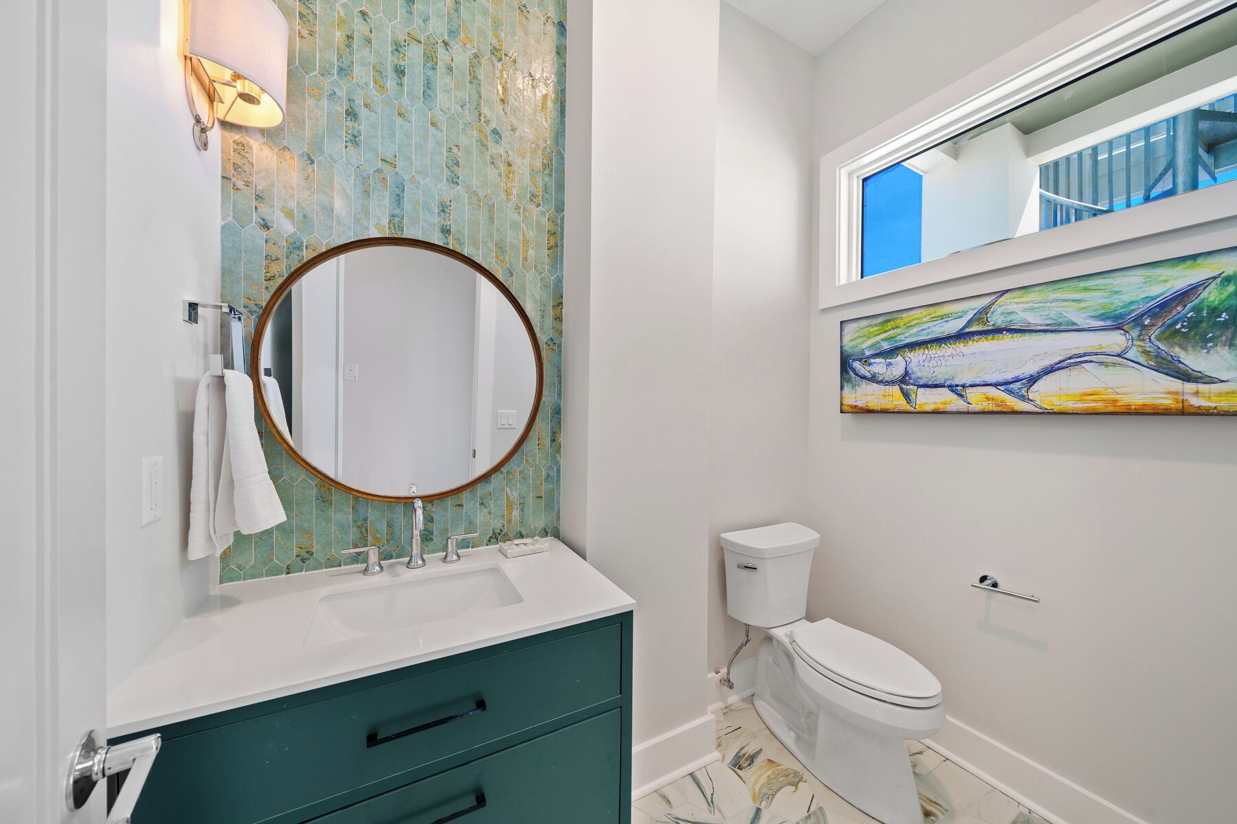 A bathroom with a white sink, a round mirror, a green cabinet, a toilet, and a large abstract painting on the wall.