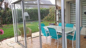 A conservatory with a glass door, a table with chairs, and a garden view. - Loue Maison Individuelle (Bénodet)
