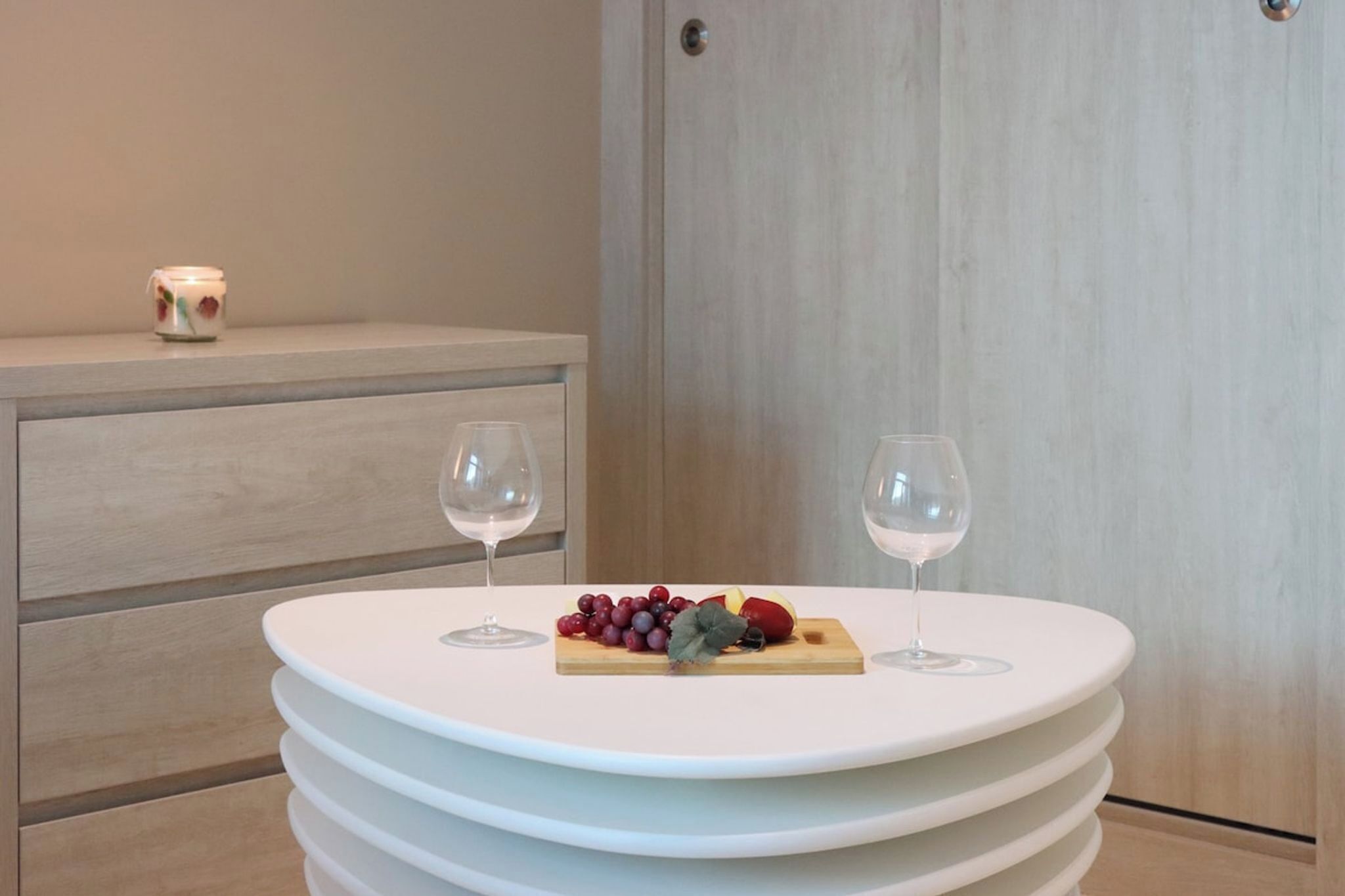 Una mesa redonda blanca con una bandeja de frutas y dos copas de vino, frente a una pared de madera con un armario al fondo.