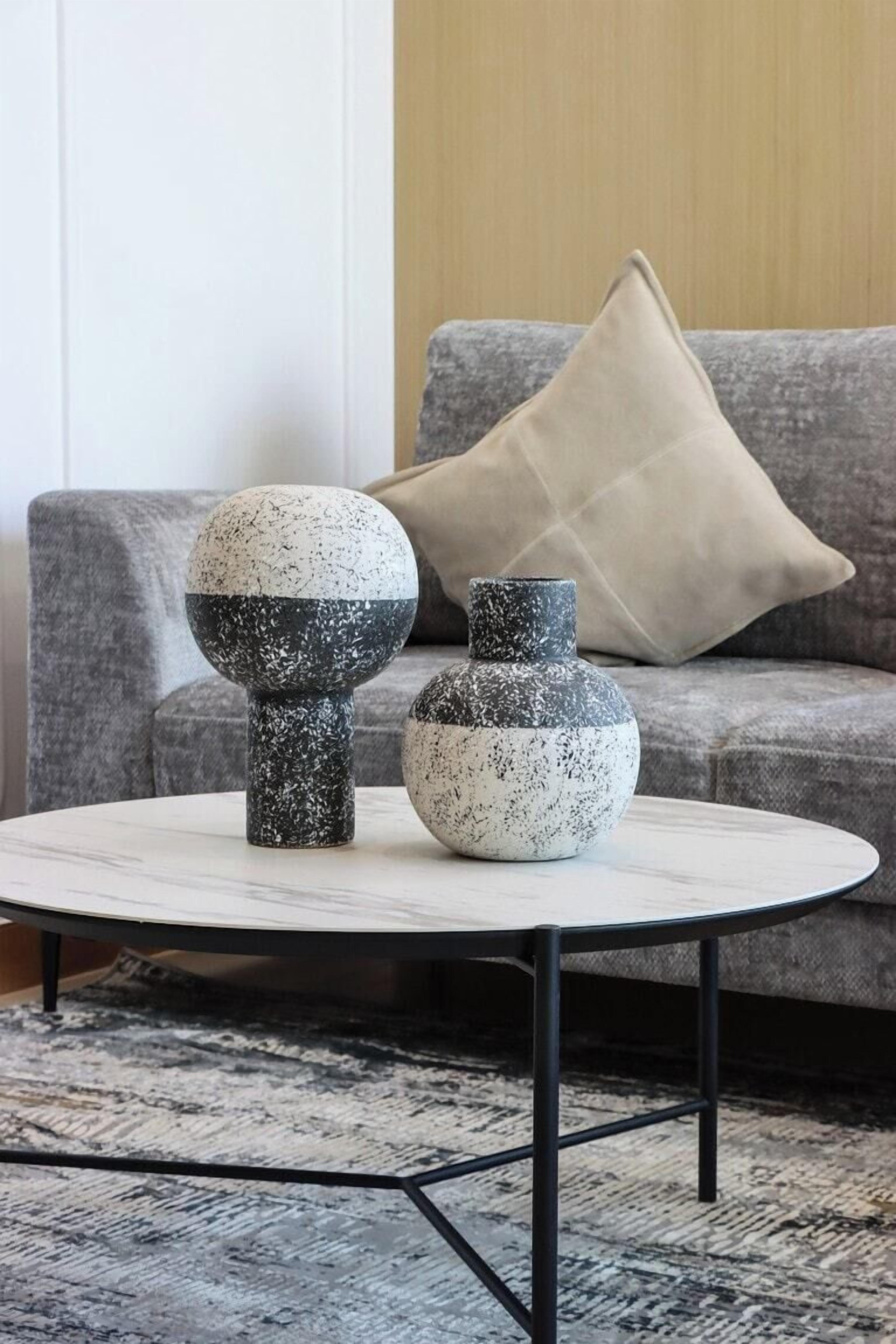 Due vasi decorati su un tavolo rotondo con un tappeto a motivi geometrici, un divano grigio con un cuscino beige e una parete rivestita in legno sullo sfondo.