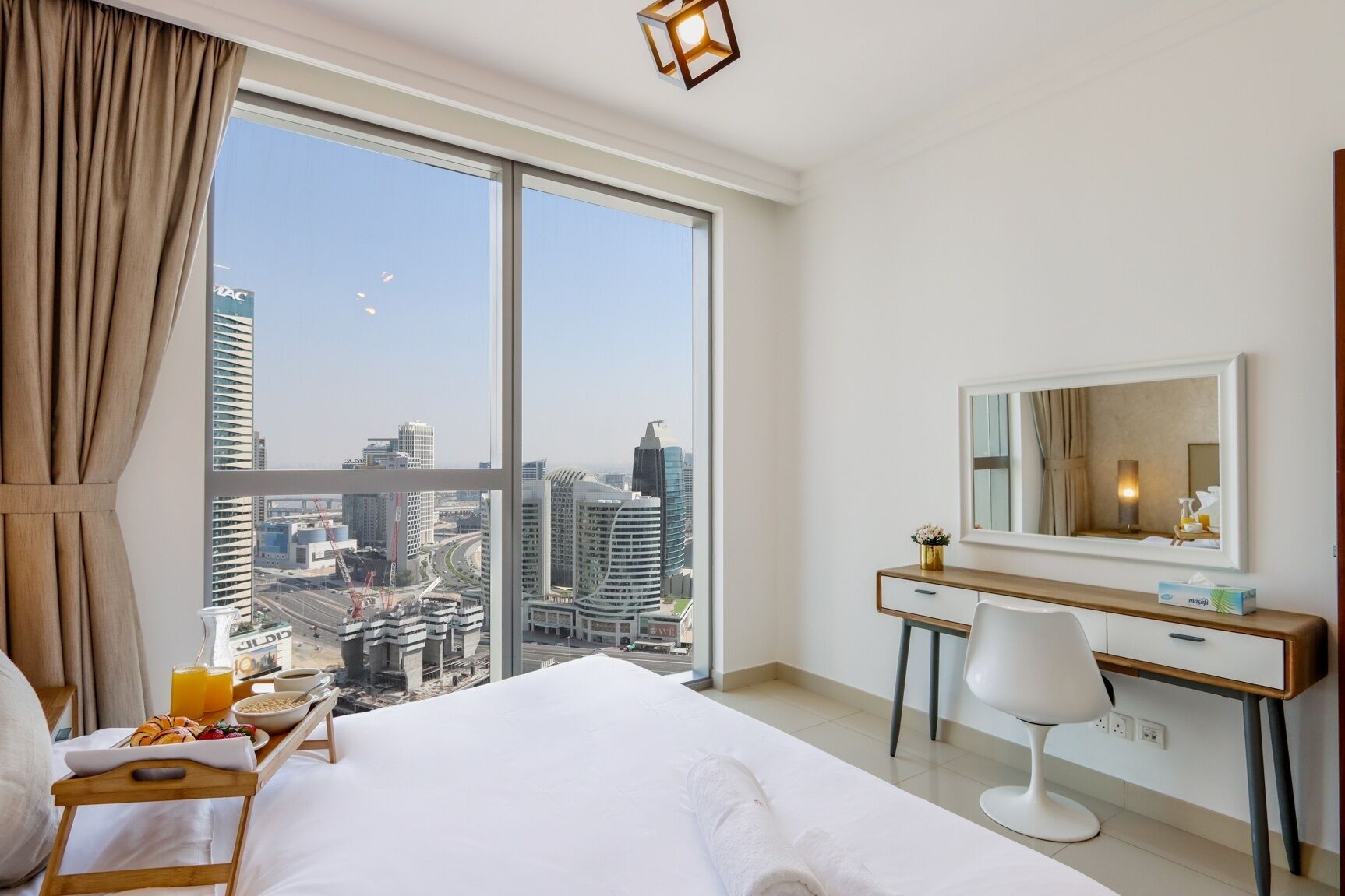 A hotel room with a large bed, a desk, a chair, and a view of the cityscape through large windows.