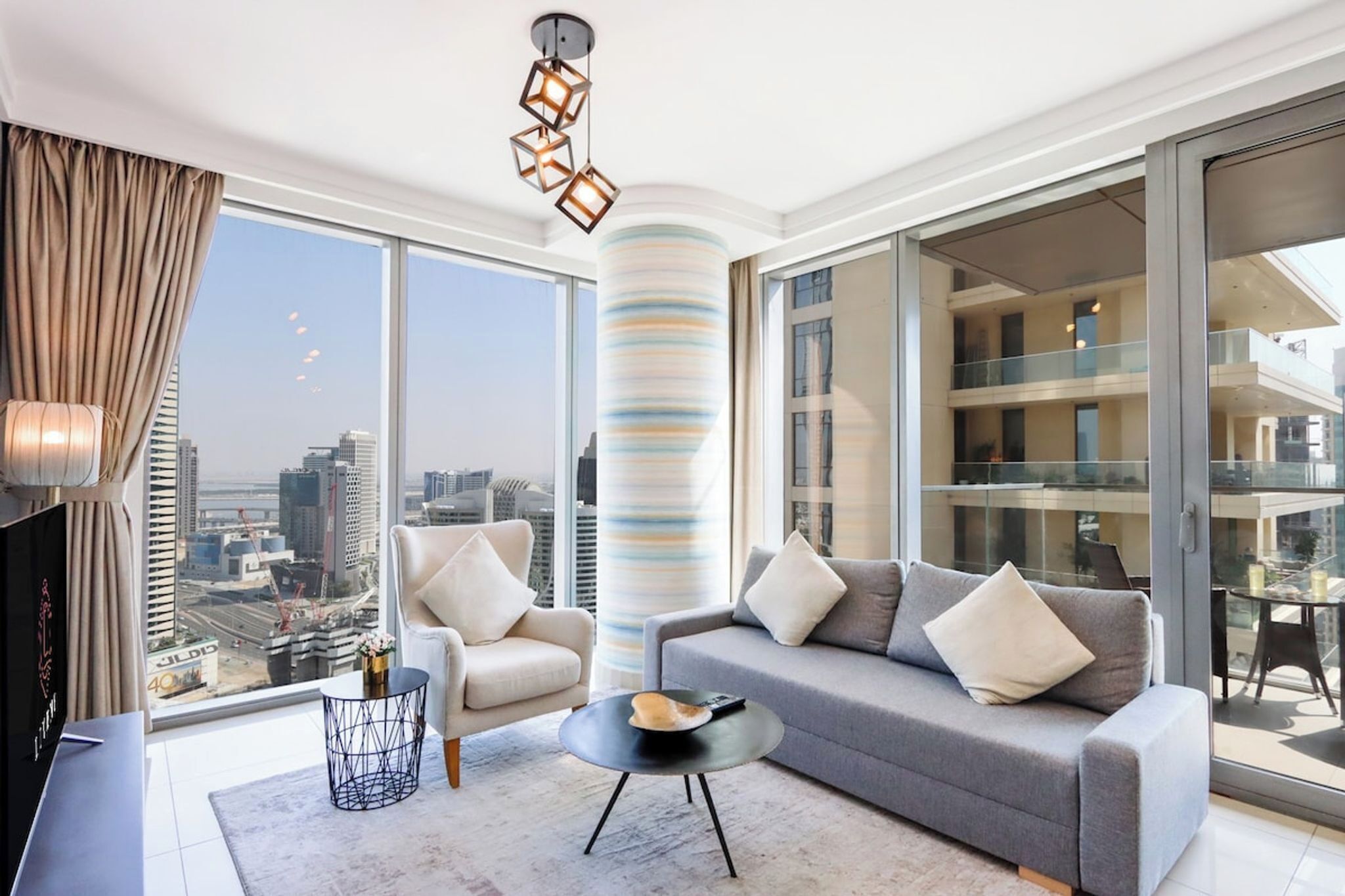 A modern living room with a sofa, armchairs, a coffee table, and a TV. A large window offers a city view.
