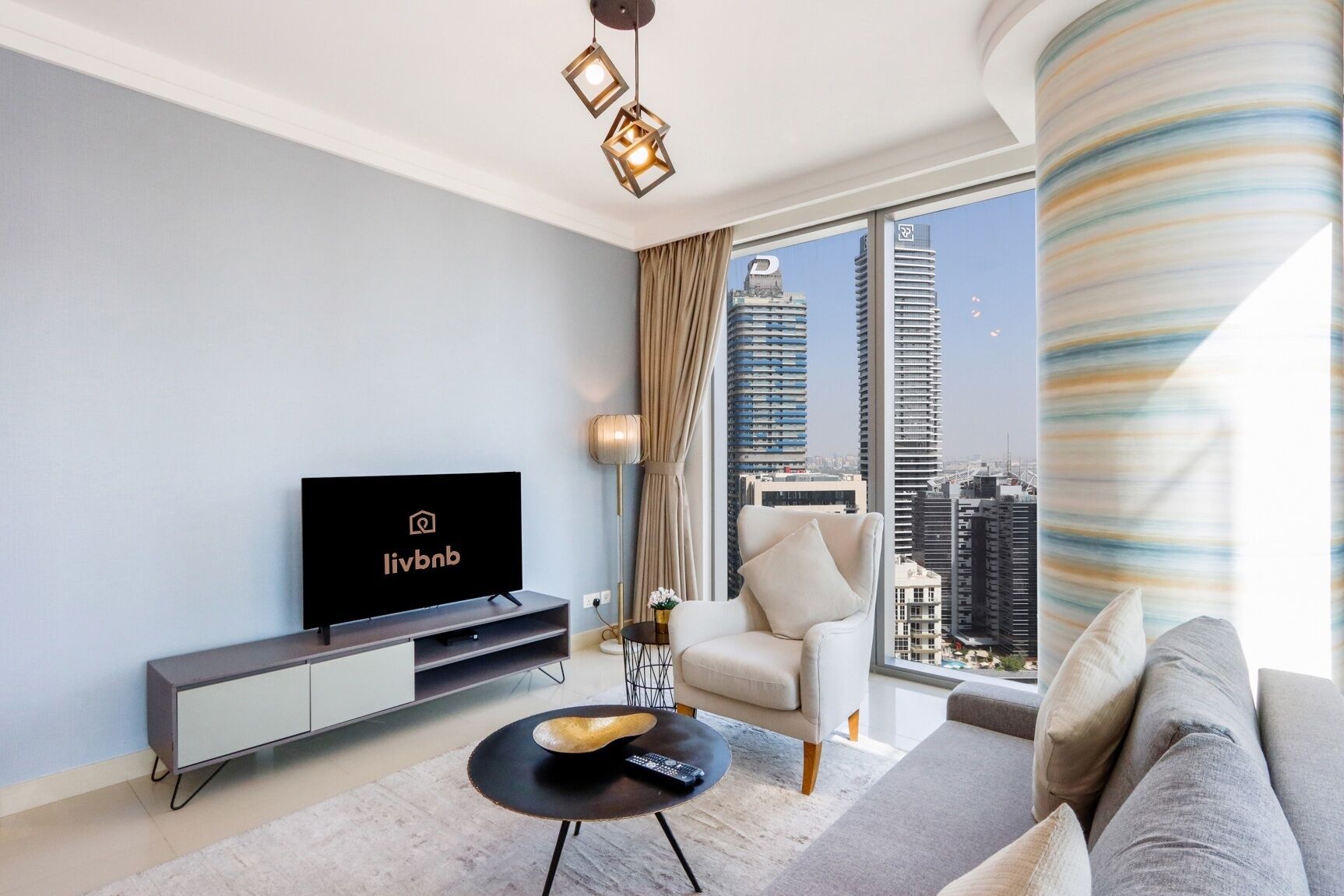 A modern living room with a flat-screen TV, a sofa, a coffee table, and a view of a cityscape through large windows.