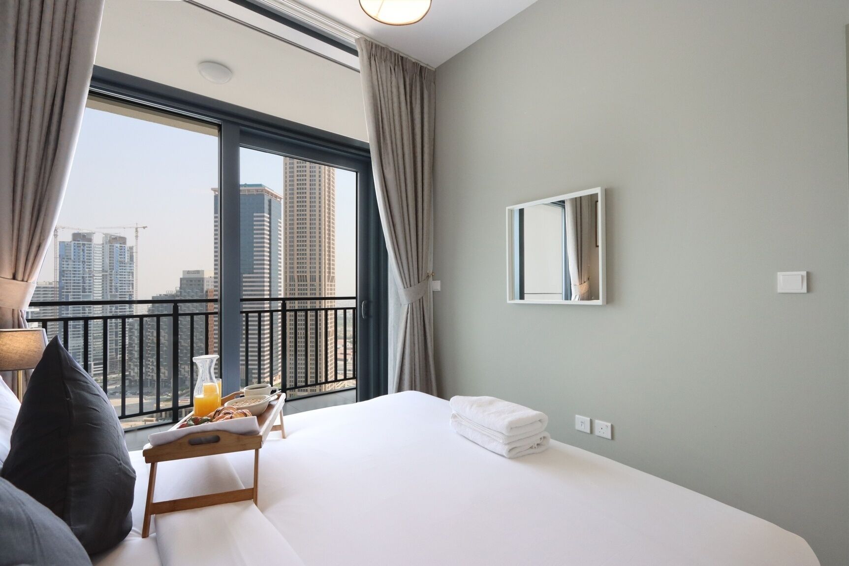 Ein Hotelzimmer mit einem großen Bett, einem Nachttisch mit Frühstücksangebot, einem Spiegel an der Wand und Blick auf Stadtgebäude durch ein großes Fenster.