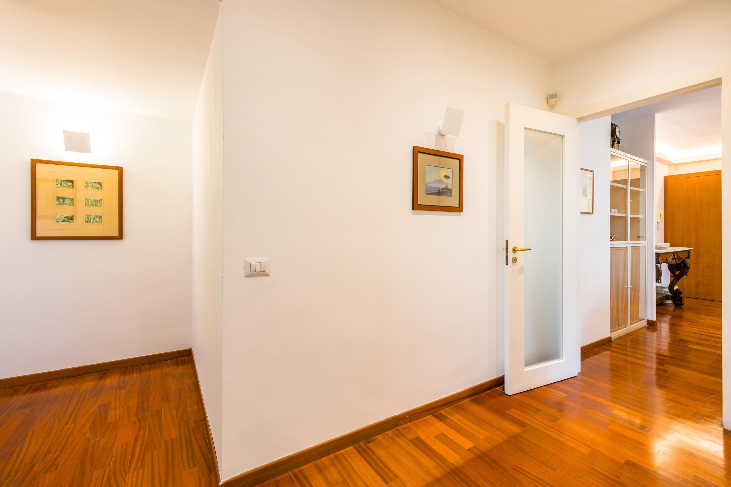 A hallway with wooden flooring, white walls, and a door leading to a room with a closet and shelves.