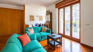 A spacious living room with a teal sofa, wooden flooring, a small table, and a large sliding glass door leading to an outdoor patio. - Colonna Classic Style Apartment by Wonderful Italy (Naples)