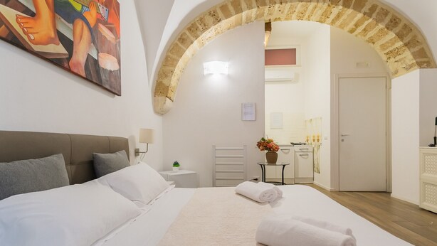 Een hotelkamer met een groot bed, een hoofdbord, een kleine tafel met een bloevas, een witte deur en een boogvormige doorgang naar een andere kamer.