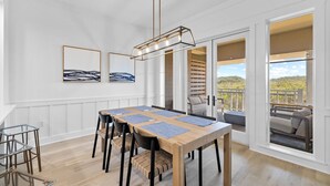 A dining area with a wooden table, black chairs, and a hanging light fixture. There are two framed artworks on the wall and a view of the outdoors through large windows. - Golf Cart | Near Private Beach Access (Watersound)
