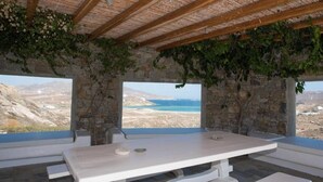 Una stanza con pareti in pietra, soffitto in legno e un tavolo con panche. Due finestre offrono vista su un paesaggio costiero.