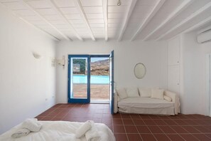 A room with a bed, a white sofa, and a view of a swimming pool through a glass door. - Mykonian Private 2 Bd Villa with Pool (Mykonos)