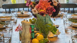 A dining table set for a meal with a vase of flowers, a bowl of citrus fruits, and glasses.