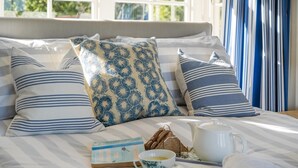 A neatly made bed with striped bedding and decorative pillows, a book, a cup of tea, and a tray with bread and a teapot.