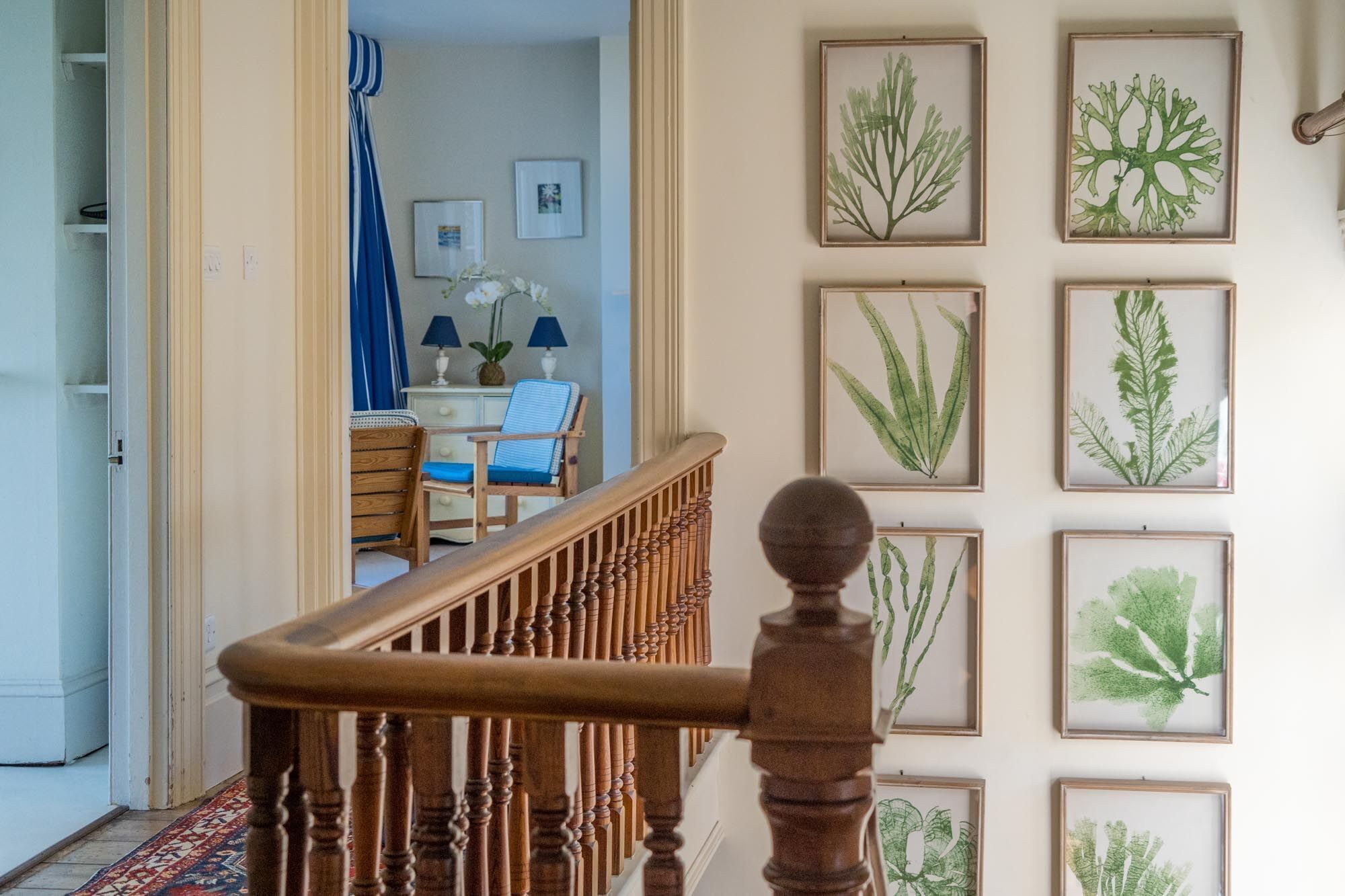 A staircase with a wooden handrail leading to a room with a dining area, a blue chair, and framed botanical prints on the wall.