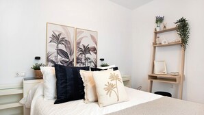 Un dormitorio ordenado con cama, almohadas, una mesita de noche, un estante en la pared con plantas y un cuadro enmarcado.