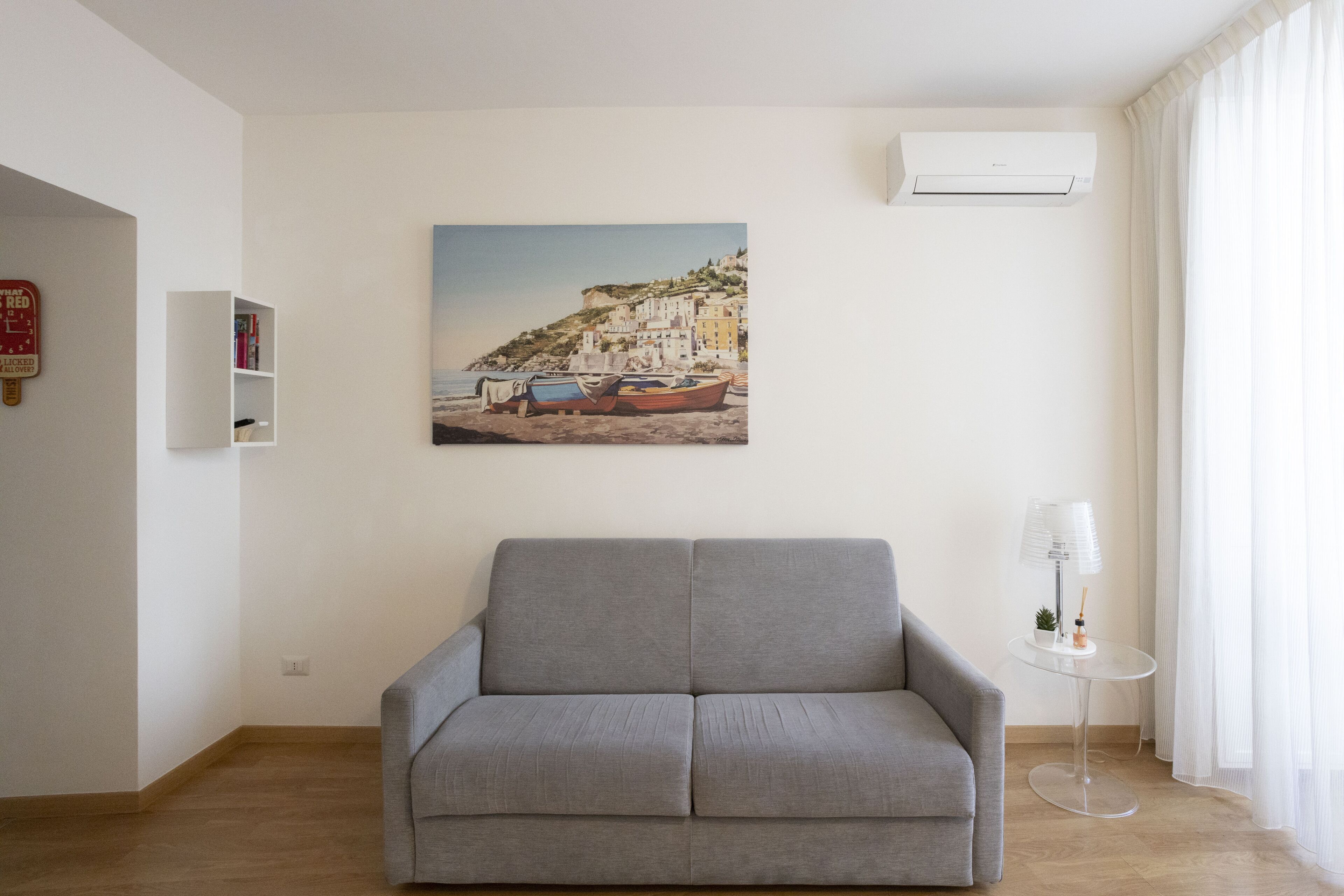 A living room with a grey sofa, a painting of boats on a cliff, a small table with a lamp, and an air conditioning unit on the wall.