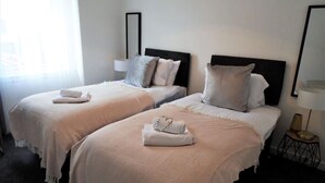 Habitación de hotel con dos camas, cada una con cubrecama y almohadas blancas, una mesita de noche con lámpara y un ventanal con cortinas translúcidas.