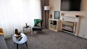 Una sala de estar con un sofá, una mesa redonda de centro, un sillón, una chimenea, un televisor montado en la pared y una estantería.