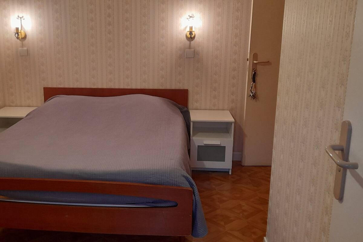 Una cama individual con estructura de madera y cubrecama gris en una habitación con papel tapiz estampado y dos lámparas de noche.