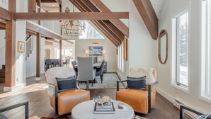 A spacious living room with a high ceiling, wooden beams, a round coffee table, leather armchairs, and a dining area in the background. - Luxurious Mt CB Home | 5 Bedroom | Home Gym | Hot Tub | Gas Grill | Mtn Views (Mt. Crested Butte)