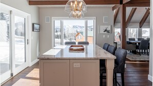 A spacious kitchen with a large island, wooden beams, and a view of mountains through large windows. - Luxurious Mt CB Home | 5 Bedroom | Home Gym | Hot Tub | Gas Grill | Mtn Views (Mt. Crested Butte)