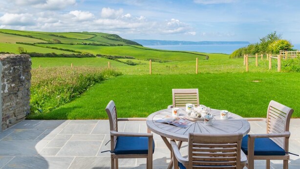 Une table ronde entourée de chaises et agrémentée d’un service à thé, placée sur une terrasse donnant sur un paysage verdoyant, avec des collines et un ciel dégagé.