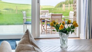 Une pièce donnant sur un champ verdoyant grâce à une porte vitrée, une table ornée d’un vase de fleurs jaunes et une paire de coussins posés sur le sol.