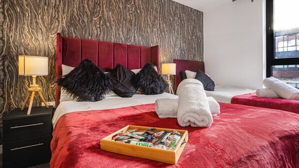 A hotel room with a red upholstered headboard, two beds, a wooden nightstand with a lamp, and a tray of snacks.
