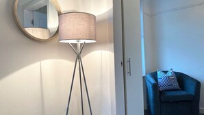 A room with a floor lamp, a round mirror, and a blue armchair with a patterned pillow.
