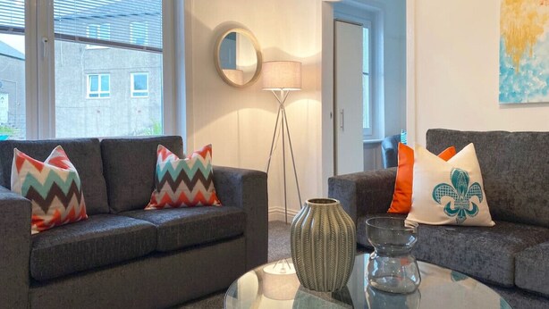 A modern living room with two grey sofas, colorful pillows, a floor lamp, a vase, and a glass table.