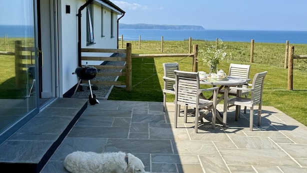 A patio with a table and chairs, a dog lying on the floor, and a view of the sea. - 2 Bed in Abbotsham (oc-g28822) (Abbotsham)