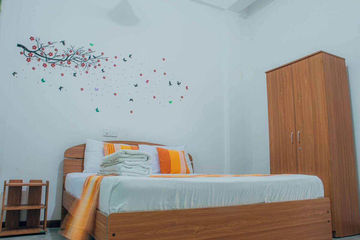 A bedroom with a wooden bed, white bedding, a wooden wardrobe, and a wall decoration of a tree with leaves and birds.