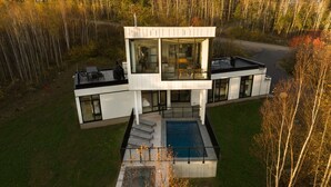 Una casa moderna, su più livelli, con un'ampia facciata vetrata, una piscina e un'area boschiva circostante.