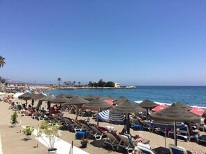 A beach with rows of sun loungers and thatched umbrellas, a clear blue sky, and a calm sea.