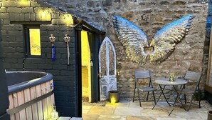 Un área de estar al aire libre de estilo rústico con una terraza de madera, un muro de piedra con un mural artístico de alas y una zona de bar con mostrador y luces de madera.
