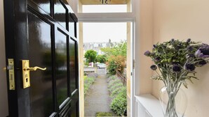 Une porte noire avec une poignée en laiton, un vase en verre transparent contenant des fleurs violettes, et une vue sur un chemin de jardin menant à une clôture blanche et à des bâtiments situés au loin.