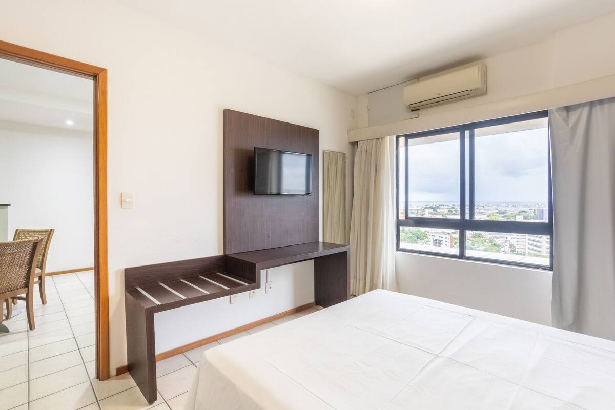 A hotel room with a bed, a TV mounted on a wooden panel, a window with a view of buildings, and a chair.